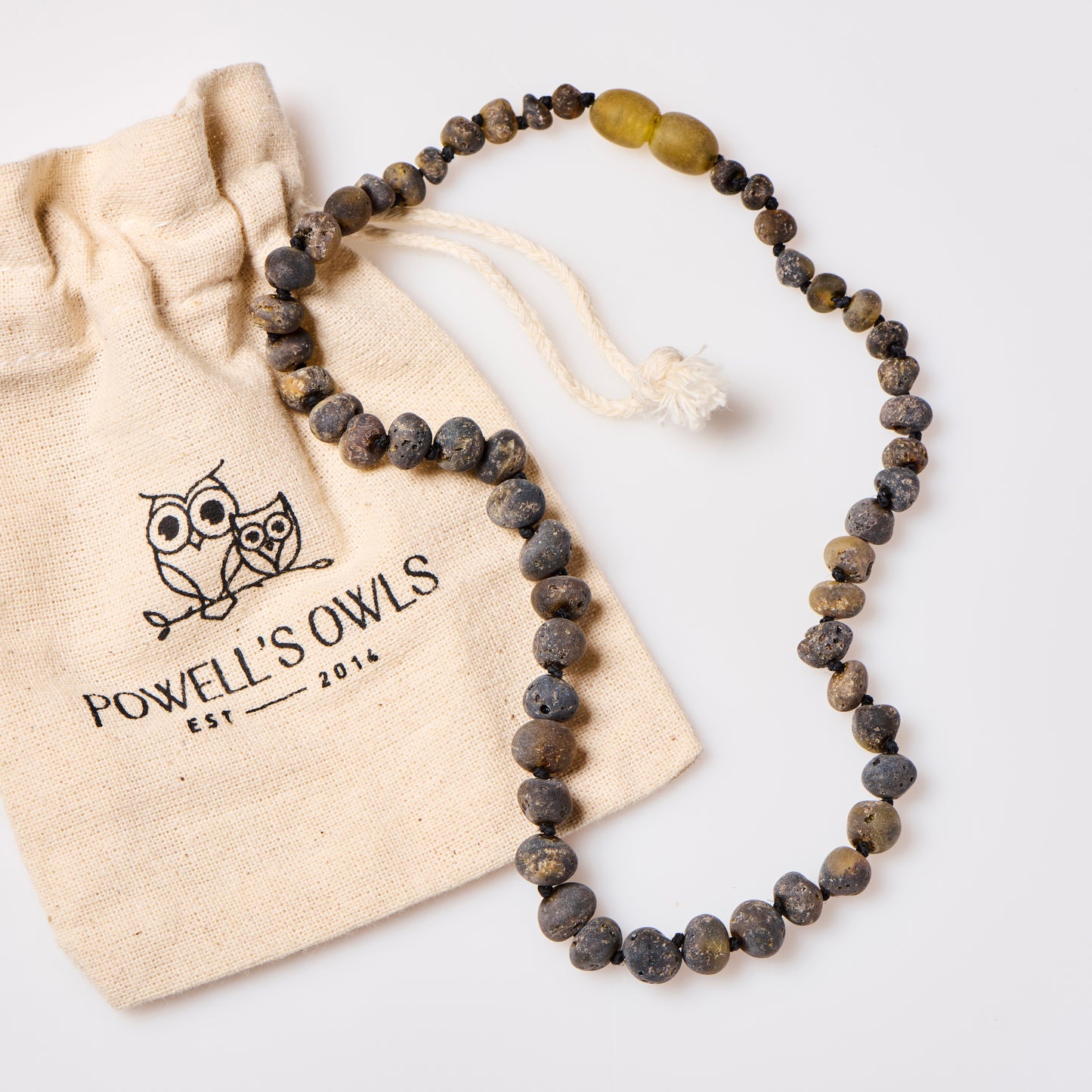 raw green amber teething necklace placed on top of a cotton jewelry pouch that says Powell's Owls